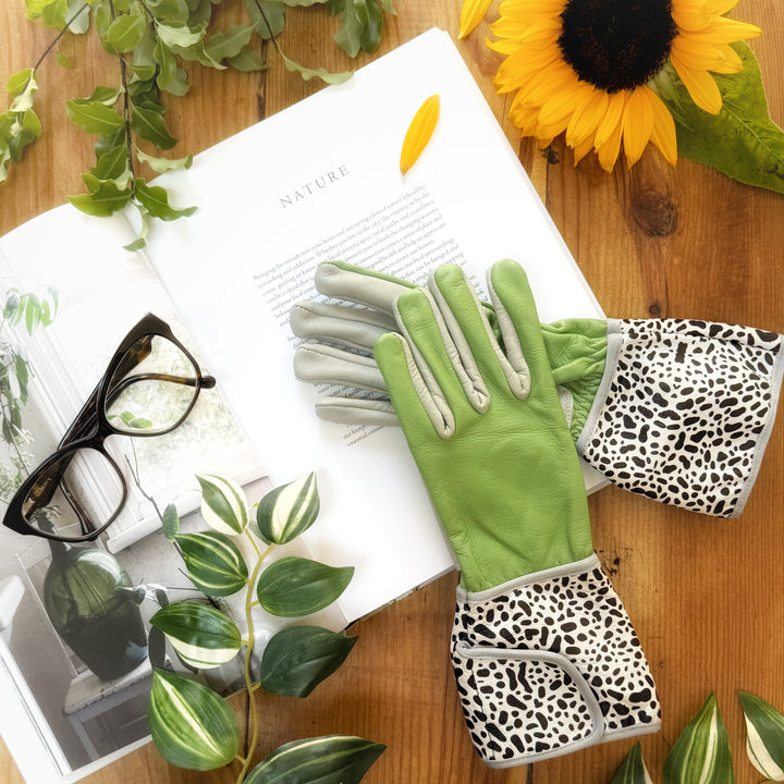 Pair of Hilda MidiGaunt sage leather gloves with leopard cuffs styled on a wooden tabletop with fresh green foliage and a sunflower.
