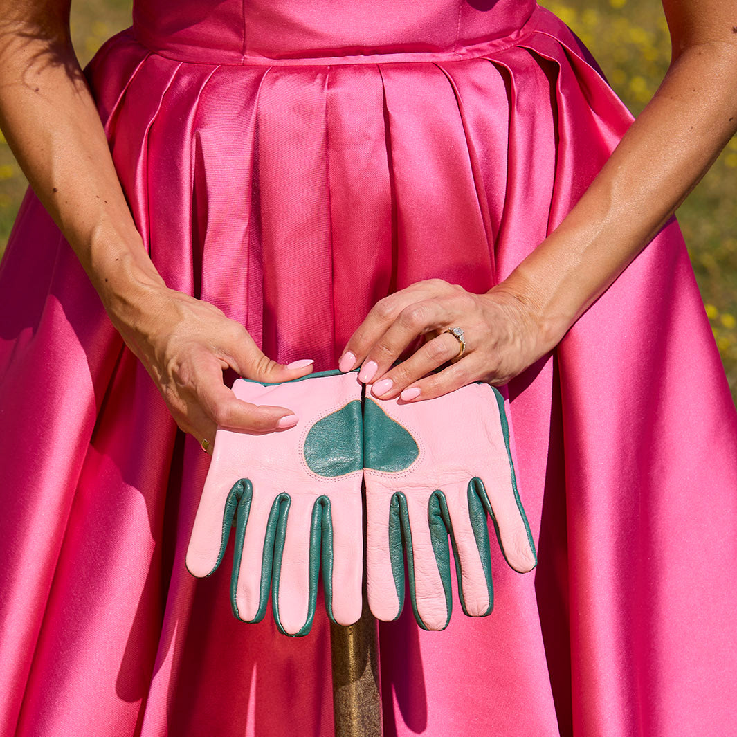 Close-up of a person holding a garden spade while wearing teal and candy pink Ivy MidiGaunt leather gardening gloves over a vibrant pink dress.
