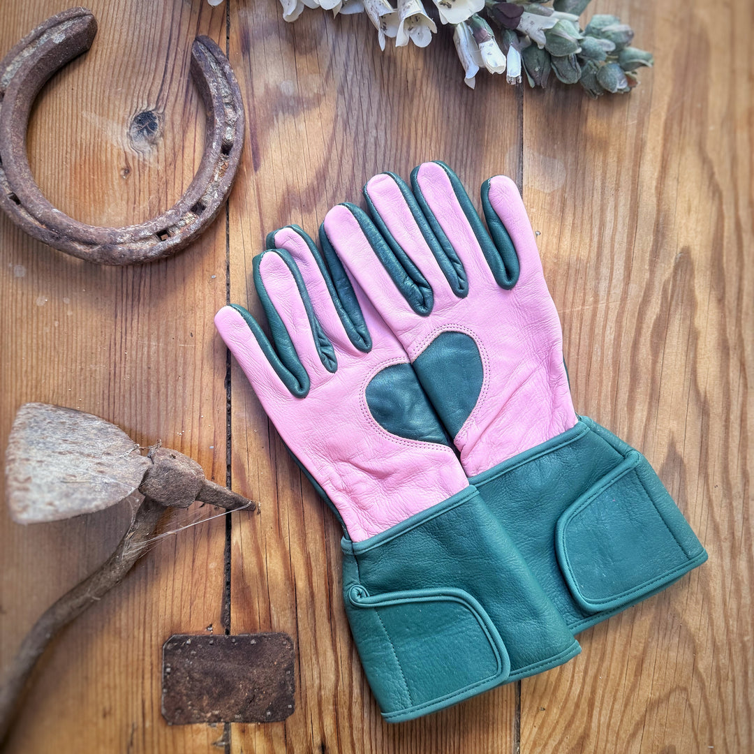 Teal and candy pink Ivy MidiGaunt gloves laid flat on a rustic wooden table surrounded by vintage garden tools and a horseshoe.