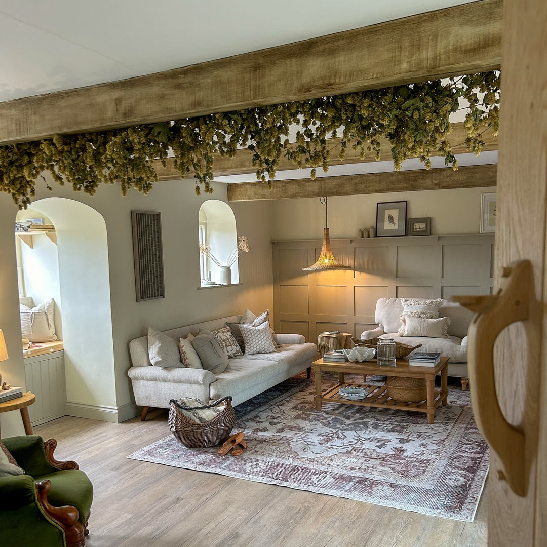 A styled living room interior featuring a cream sofa, a wooden coffee table, a patterned area rug, light wood flooring, arched windows, and a rustic wooden ceiling beam decorated with green ivy.