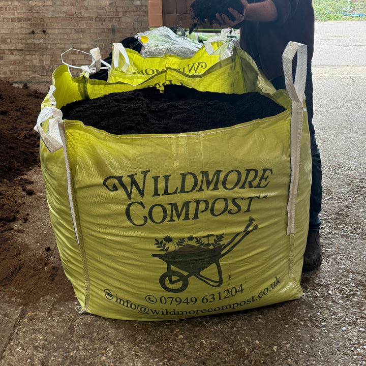 Person holding a bag of Wildmore Compost on a concrete surface with a building in the background.