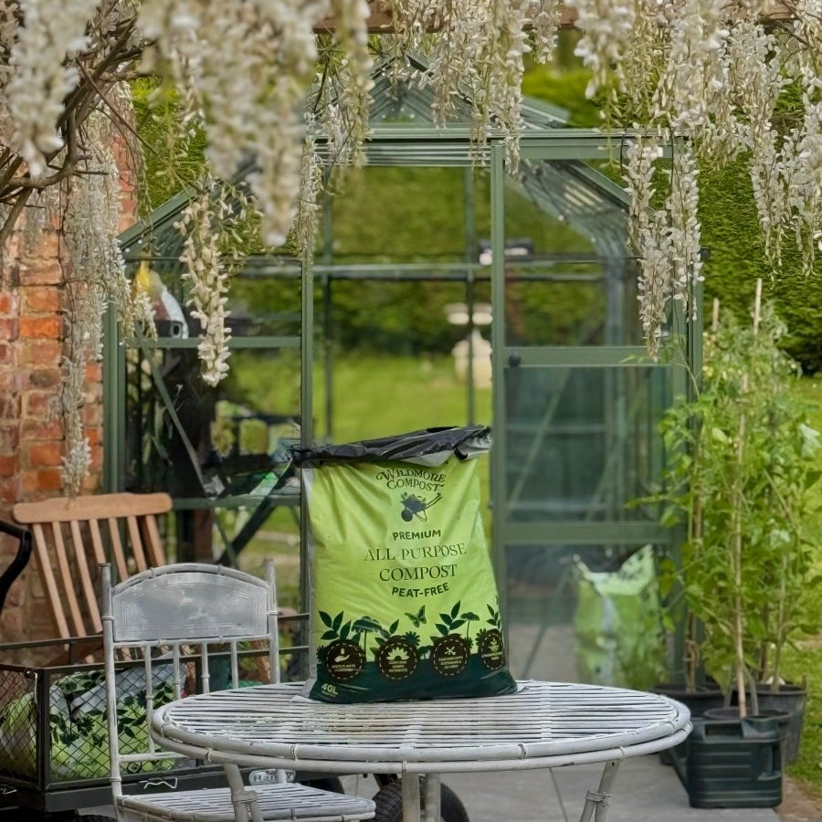 Garden setting with a table, chairs, and a compost bag in front of a greenhouse.