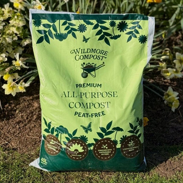 A 40L bag of Wildmore Compost Premium All Purpose Peat-Free compost, standing upright on grass in a garden with daffodils in the background.