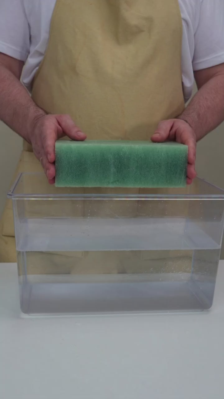 Hands gently submerging and compressing a green Phoam compostable floral foam block underwater in a clear container to absorb water.