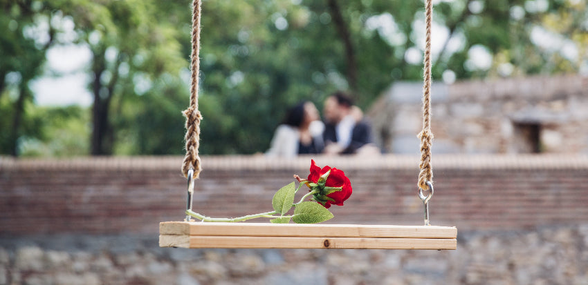 A wooden swing suspended by ropes outdoors, holding a single red rose with green leaves, with a blurred couple in the background against trees and a stone wall.