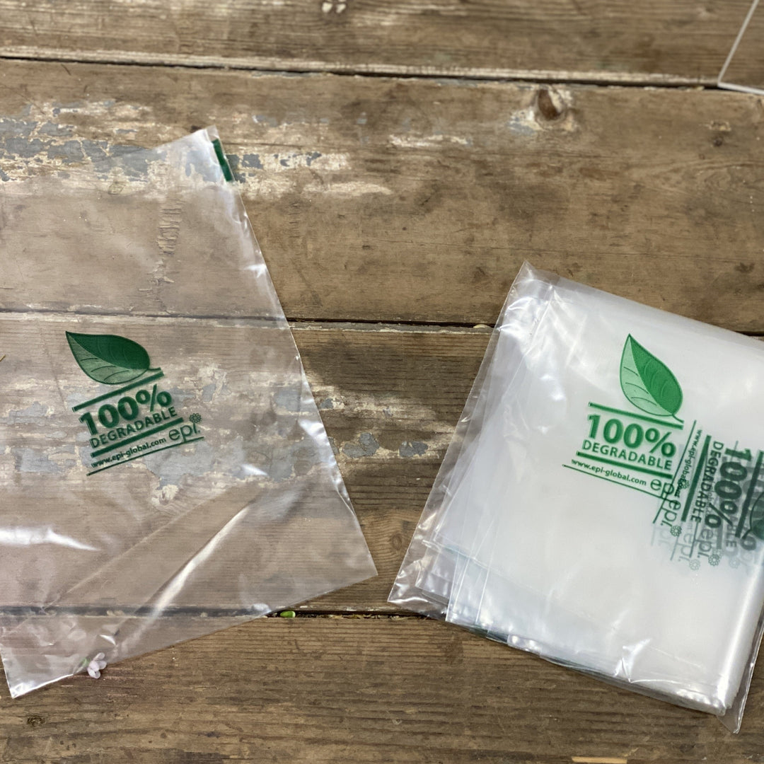 Two transparent biodegradable plastic bags with green leaf logos and text indicating 100% biodegradable on a wooden surface.