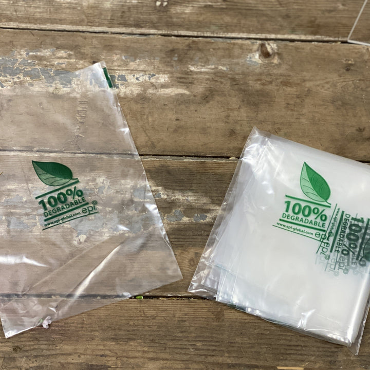 Two transparent biodegradable plastic bags with green leaf logos and text indicating 100% biodegradable on a wooden surface.