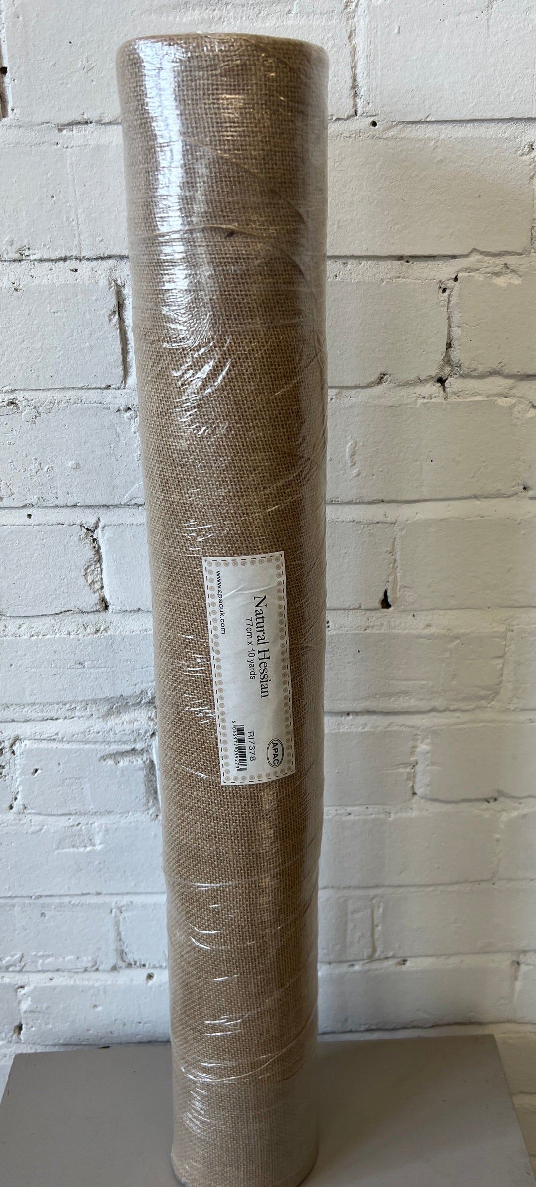 A roll of natural color hessian fabric, 77cm wide, displayed against a white brick wall background.