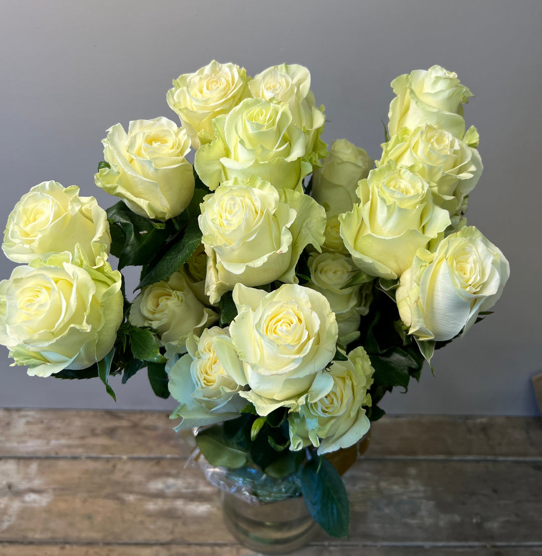 A bouquet of white roses with green leaves, arranged in a clear vase, placed on a wooden surface.