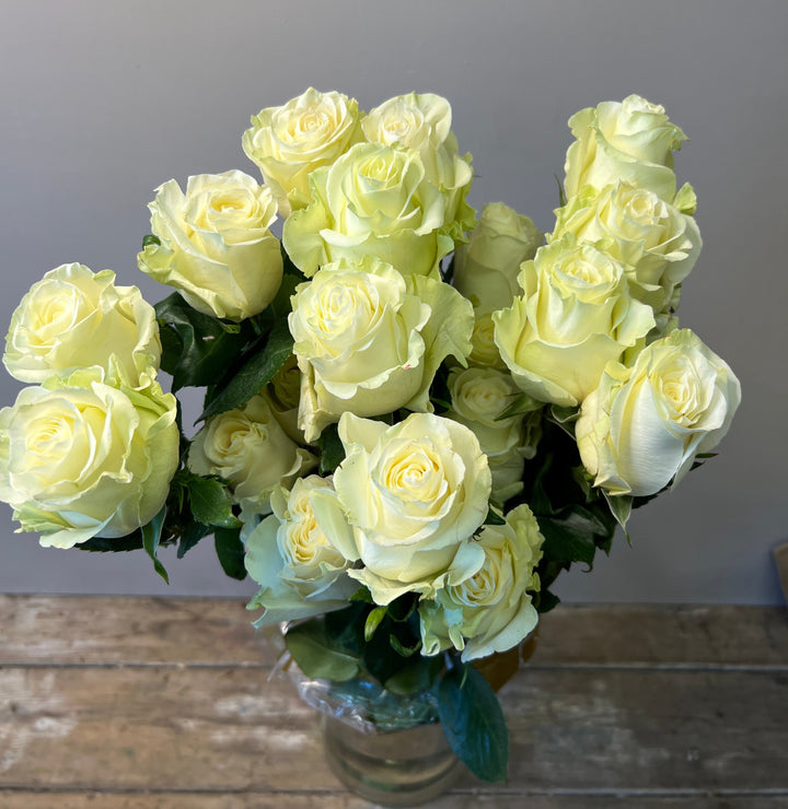 A bouquet of white roses with green leaves, arranged in a clear vase, placed on a wooden surface.